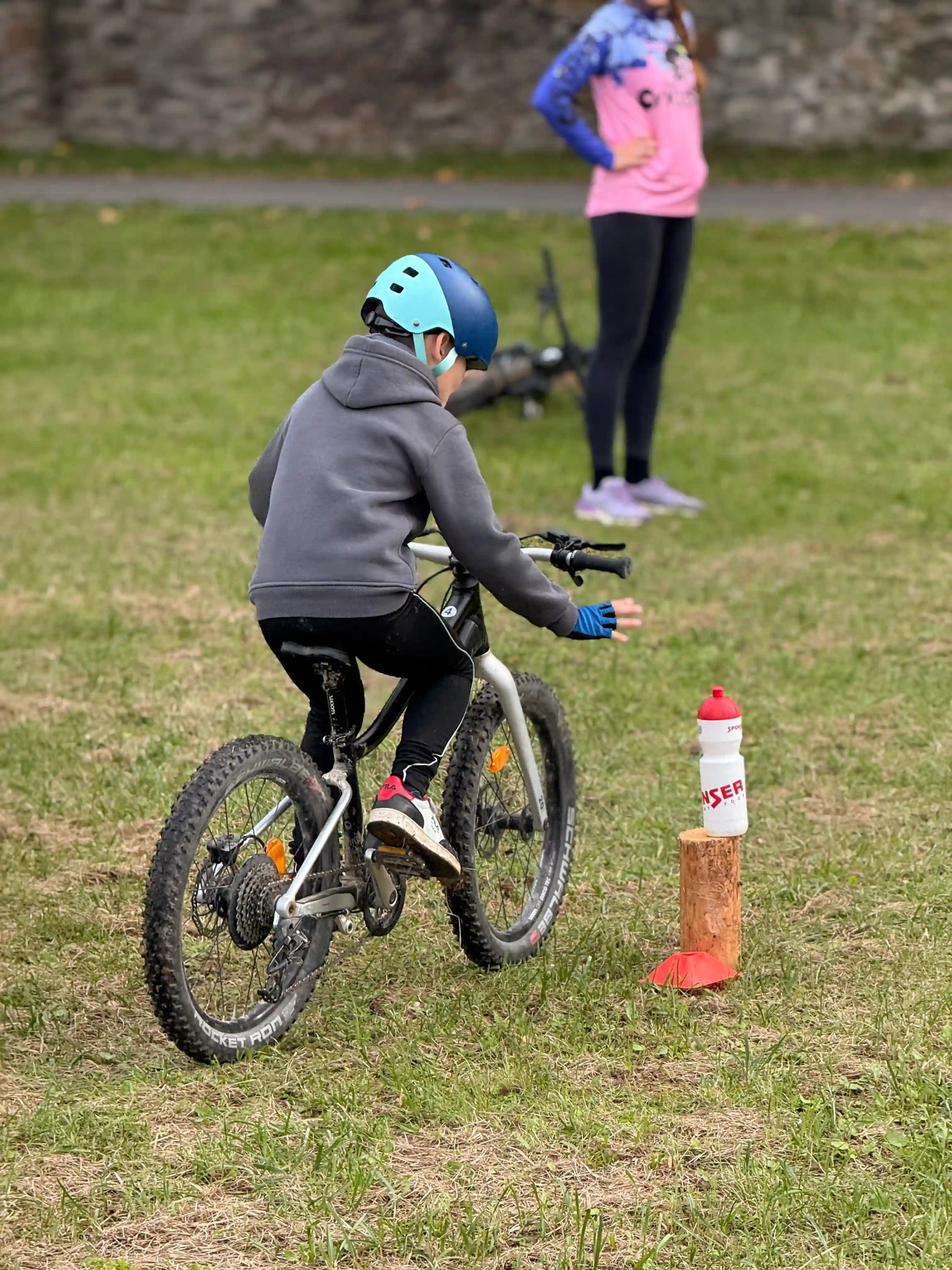 Dítě jede na kole a snaží za jízdy vzít flašku z pařezu.